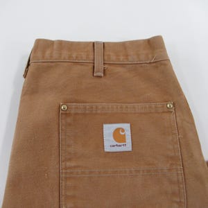 Puede incluir: Pantal&oacute;n de trabajo marr&oacute;n con un gran bolsillo de parche en el lado derecho. El bolsillo tiene un parche de logotipo de Carhartt con el texto "carhartt" en blanco.
