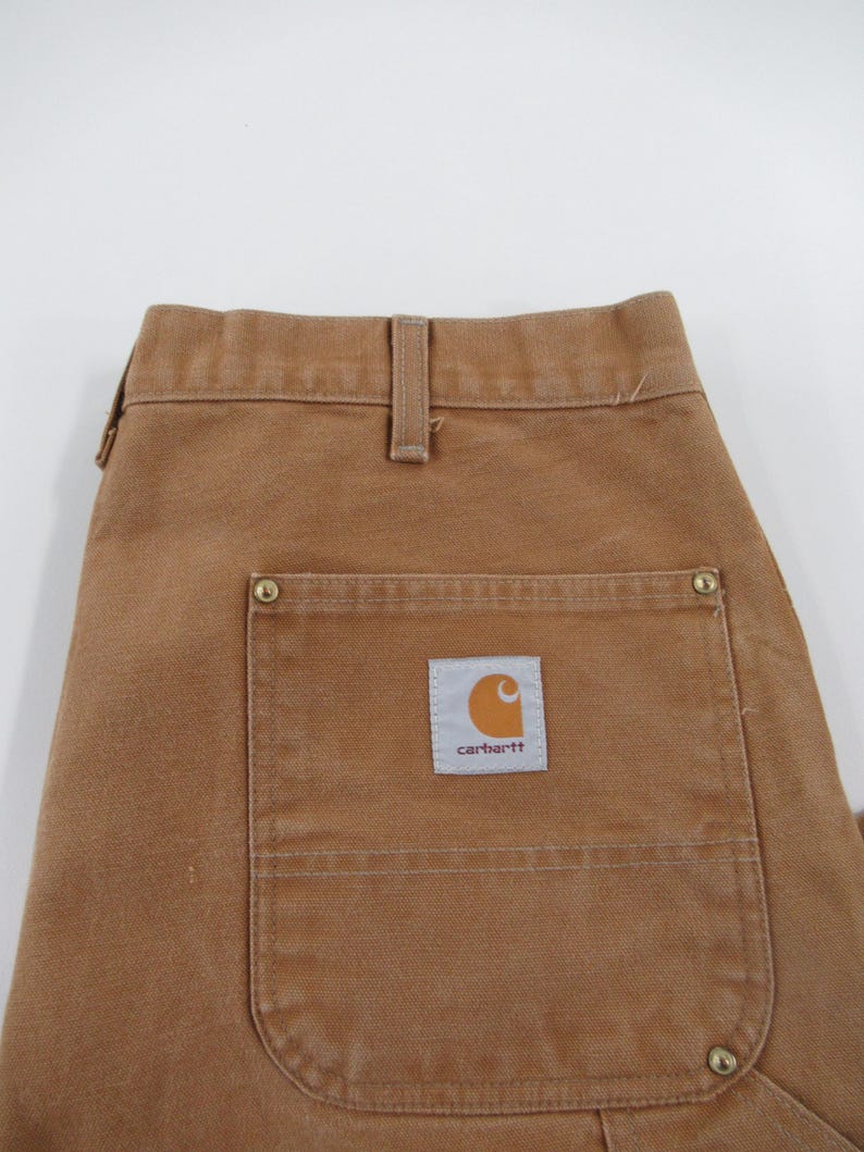 Puede incluir: Pantal&oacute;n de trabajo marr&oacute;n con un gran bolsillo de parche en el lado derecho. El bolsillo tiene un parche de logotipo de Carhartt con el texto "carhartt" en blanco.