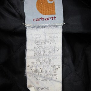 May include: A white Carhartt clothing label with black text and a red logo. The label includes care instructions and size information: "MACHINE WASH WARM SEPARATELY. DO NOT BLEACH. TUMBLE DRY MEDIUM. REMOVE PROMPTLY. WHEN DESIRED, ONLY IRON SHELL SIDE. SHELL: 100% COTTON LINING: 100% NYLON QUILTED 100% POLYESTER BATT ASSEMBLED IN MEXICO SEE REVERS FOR WARNING X-LARGE 178CMT"