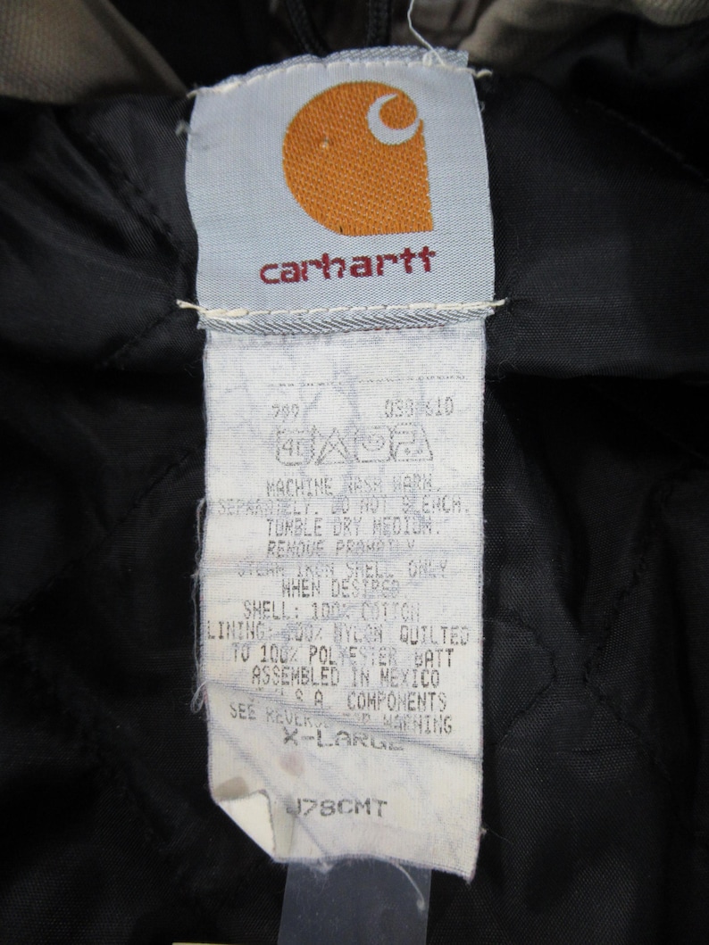 May include: A white Carhartt clothing label with black text and a red logo. The label includes care instructions and size information: "MACHINE WASH WARM SEPARATELY. DO NOT BLEACH. TUMBLE DRY MEDIUM. REMOVE PROMPTLY. WHEN DESIRED, ONLY IRON SHELL SIDE. SHELL: 100% COTTON LINING: 100% NYLON QUILTED 100% POLYESTER BATT ASSEMBLED IN MEXICO SEE REVERS FOR WARNING X-LARGE 178CMT"