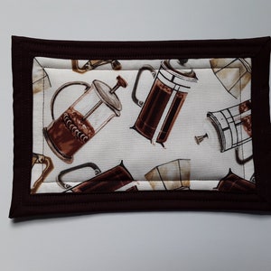 May include: Rectangular coffee-themed padded placemat. The placemat features a repeating pattern of brown French presses on a cream background. A dark brown border frames the design, adding a contrasting edge to the coffee-themed accessory.