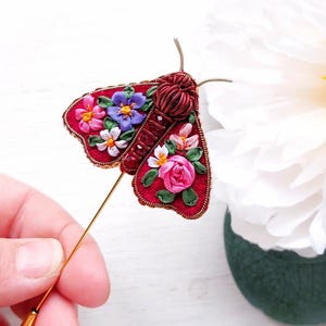 May include: A handcrafted, red moth brooch with floral embroidery in pink, purple, and white. The brooch has a gold pin and antenna details. The design is held by a hand, with a white flower in the background.