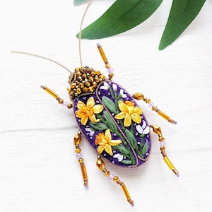 May include: A handcrafted brooch in the shape of a beetle. The brooch features a purple body with yellow flower and green leaf embroidery. The head is covered in gold beads, and the legs are gold with purple accents.