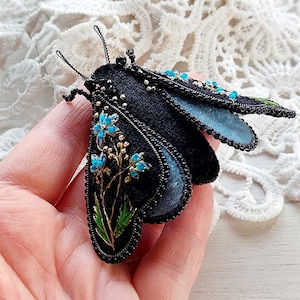 Op de afbeelding: Zwarte fluwelen motten broche met blauwe en gouden geborduurde bloemen. De mot heeft een driedimensionaal ontwerp en is versierd met zwarte kralen.