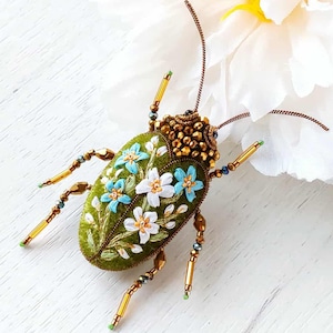 May include: A handcrafted, green velvet beetle brooch with embroidered white and blue flowers. The beetle's head is adorned with gold beads, and its legs and antennae are made of gold-colored wire and beads. A white flower is in the background.