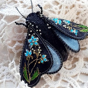 May include: A black and blue embroidered moth brooch with a floral design featuring blue flowers and green leaves. The brooch is decorated with silver beads and has a black felt backing.