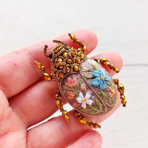 May include: A detailed, embroidered beetle brooch. The body is a light brown with floral embroidery in pink, blue, and white. The head and legs are adorned with gold-colored beads. A unique, handcrafted accessory.