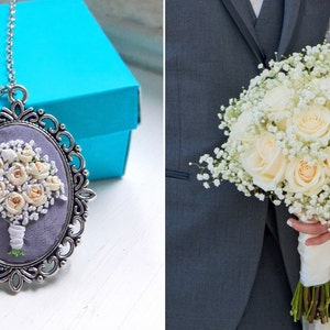 May include: A silver pendant necklace with a white and cream floral bouquet embroidered on a gray background. The bouquet is in a silver oval frame with a decorative border. The necklace is on a blue box.