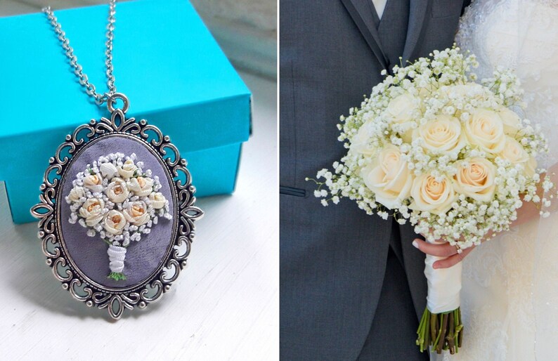 May include: A silver pendant necklace with a white and cream floral bouquet embroidered on a gray background. The bouquet is in a silver oval frame with a decorative border. The necklace is on a blue box.
