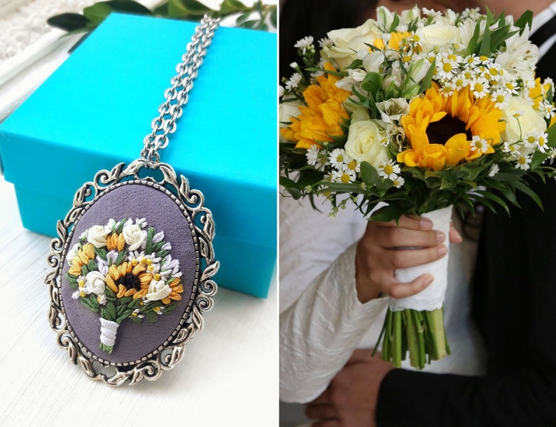 May include: A silver necklace with a round pendant featuring an embroidered bouquet of white and yellow flowers, including sunflowers, on a gray background.
