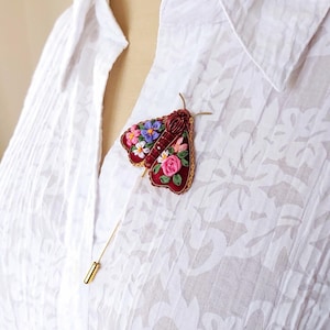 May include: A burgundy embroidered moth brooch with floral details in pink, purple, and white. The brooch is attached to a white button-down shirt with a gold pin.