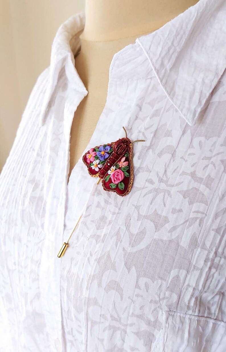 May include: A burgundy embroidered moth brooch with floral details in pink, purple, and white. The brooch is attached to a white button-down shirt with a gold pin.