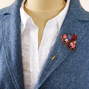 May include: A blue blazer with a white shirt and a decorative moth brooch. The brooch is embroidered with colorful flowers and has a gold pin. A gold stick pin is also visible.