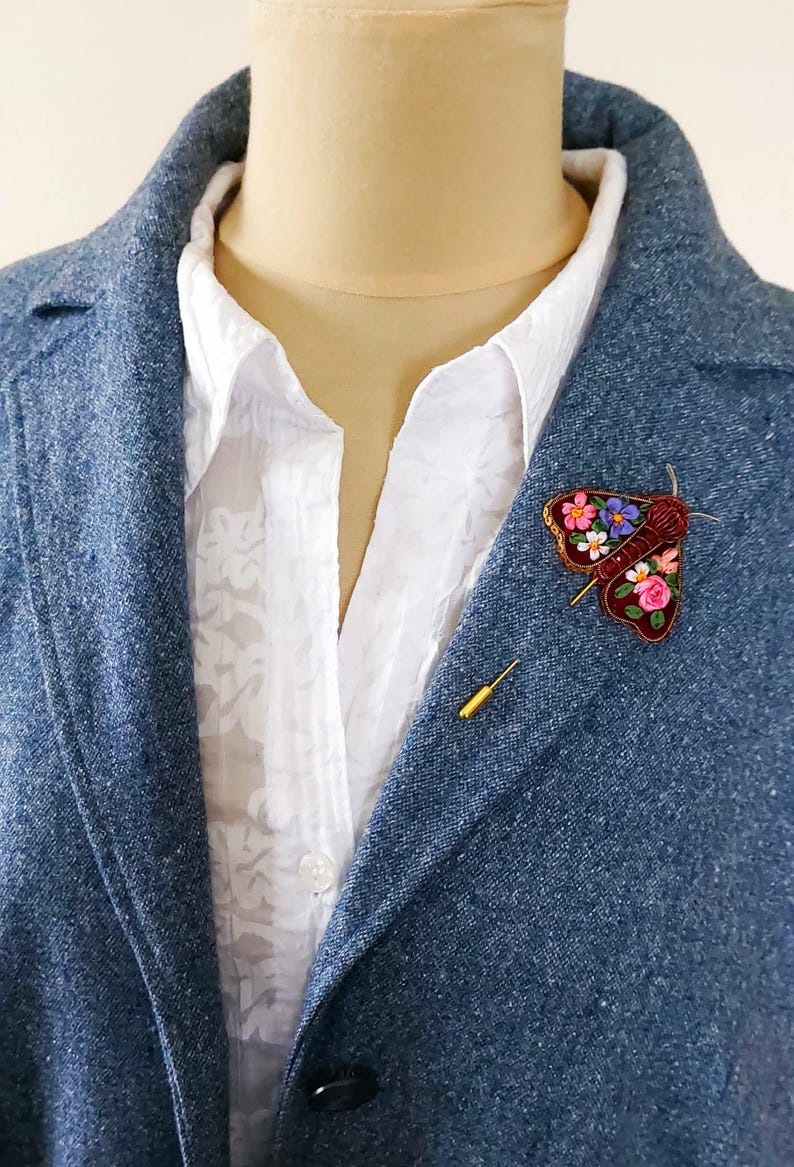 May include: A blue blazer with a white shirt and a decorative moth brooch. The brooch is embroidered with colorful flowers and has a gold pin. A gold stick pin is also visible.