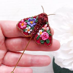May include: A handcrafted red moth brooch with floral embroidery. The brooch features a gold pin and is adorned with colorful flowers, including pink roses, white daisies, and purple blooms. The moth's body is made of twisted red wire.