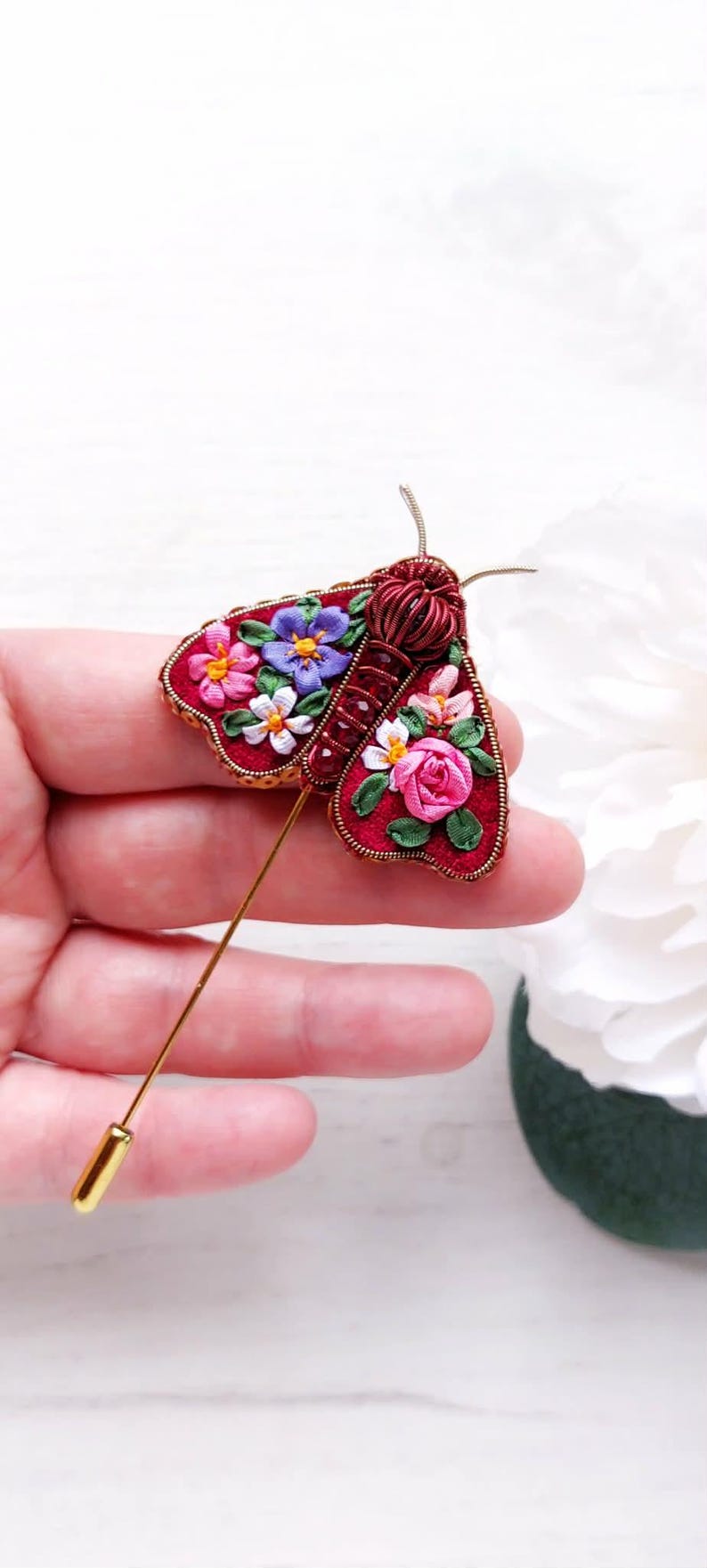 May include: A handcrafted red moth brooch with floral embroidery. The brooch features a gold pin and is adorned with colorful flowers, including pink roses, white daisies, and purple blooms. The moth's body is made of twisted red wire.