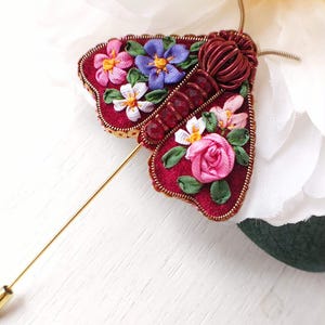 May include: A handcrafted burgundy moth brooch with floral embroidery. The brooch features a gold pin and is adorned with pink, purple, and white flowers. The moth's body is made of coiled wire.