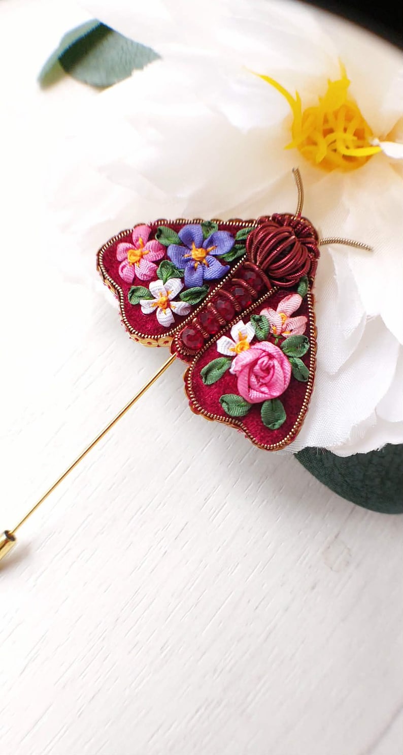May include: A handcrafted burgundy moth brooch with floral embroidery. The brooch features a gold pin and is adorned with pink, purple, and white flowers. The moth's body is made of coiled wire.