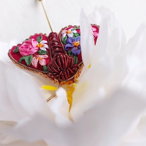 May include: A handcrafted, red fabric butterfly brooch with intricate floral embroidery in pink, purple, and white. The brooch features a gold-colored metal frame and pin, set against a white background.