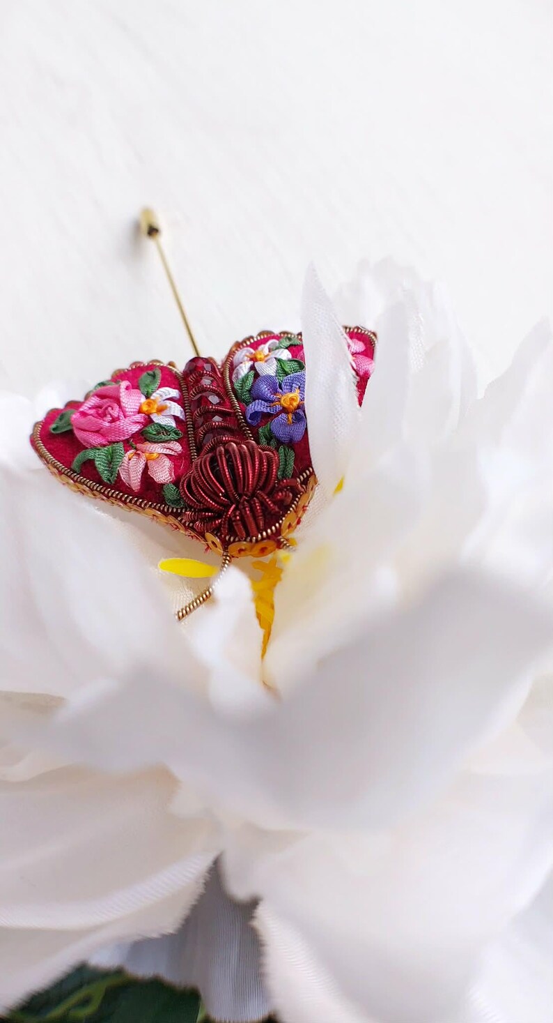 May include: A handcrafted, red fabric butterfly brooch with intricate floral embroidery in pink, purple, and white. The brooch features a gold-colored metal frame and pin, set against a white background.