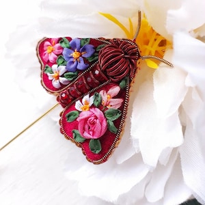 May include: A handcrafted, red fabric moth brooch with embroidered floral details. The moth's wings feature pink, purple, and white flowers, with a gold-toned pin. The brooch is set against a white flower.