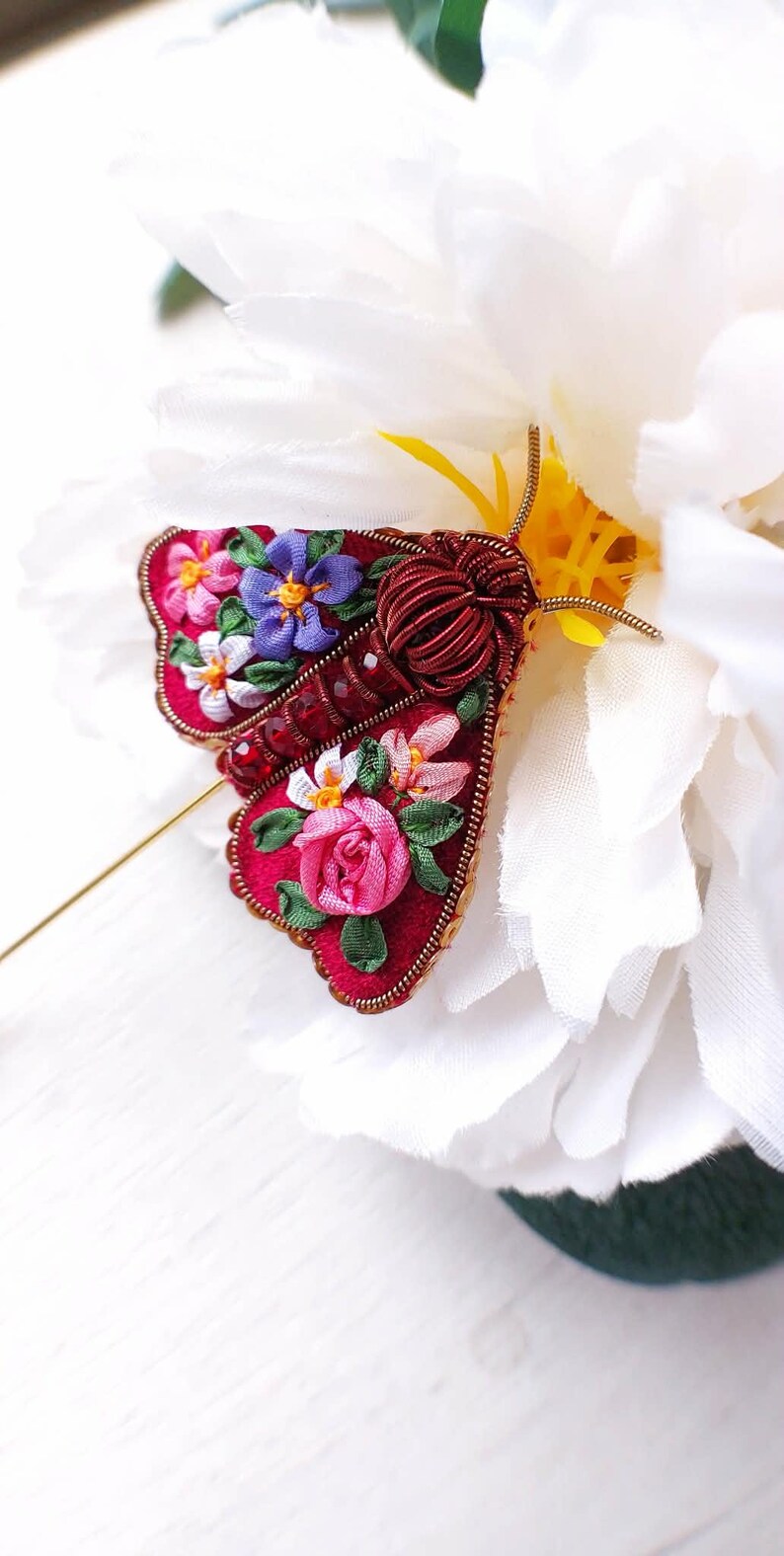 May include: A handcrafted, red fabric moth brooch with embroidered floral details. The moth's wings feature pink, purple, and white flowers, with a gold-toned pin. The brooch is set against a white flower.