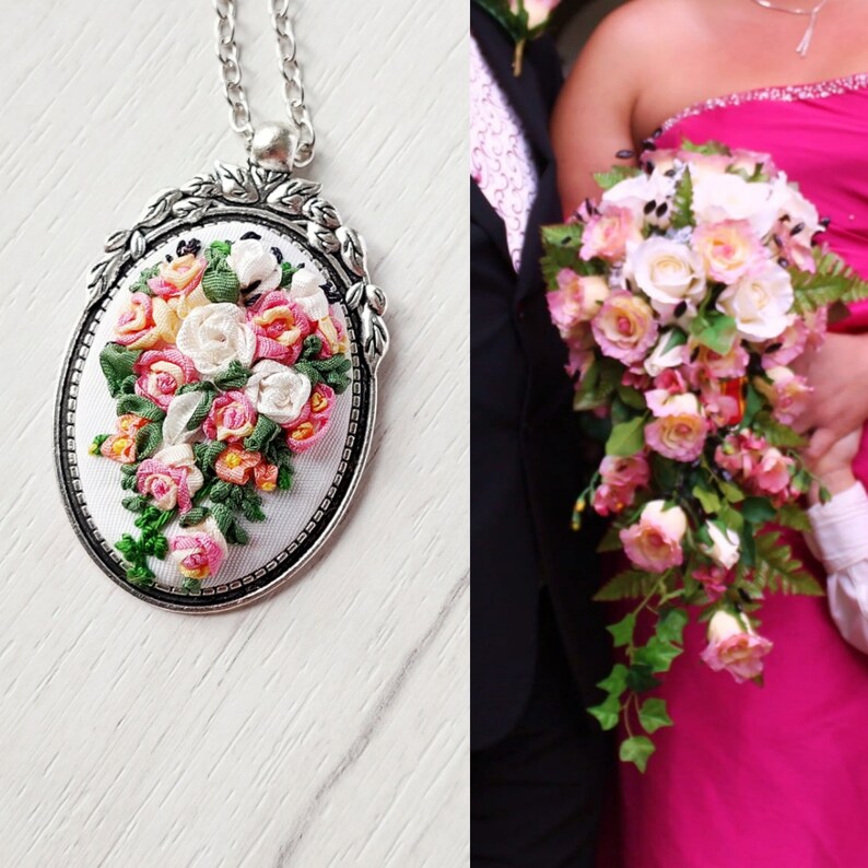 May include: A silver oval pendant necklace with a white satin background and a pink and white floral design. The pendant features a bouquet of pink and white roses, with green leaves and stems.