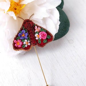 May include: A handcrafted, red moth brooch with embroidered floral details in pink, purple, and white. The brooch has a gold pin and is set against a white flower and green leaves.