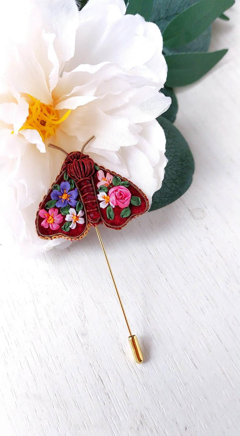 May include: A handcrafted, red moth brooch with embroidered floral details in pink, purple, and white. The brooch has a gold pin and is set against a white flower and green leaves.