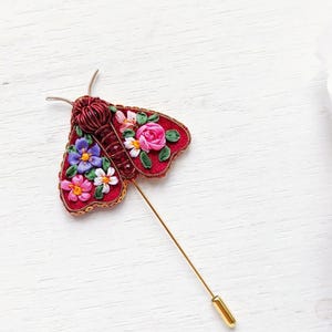 May include: A handcrafted red moth brooch with floral embroidery in pink, purple, and white. The brooch has a gold pin and is set against a white wooden background. The moth's body is a deep red, and the wings are adorned with colorful flowers.
