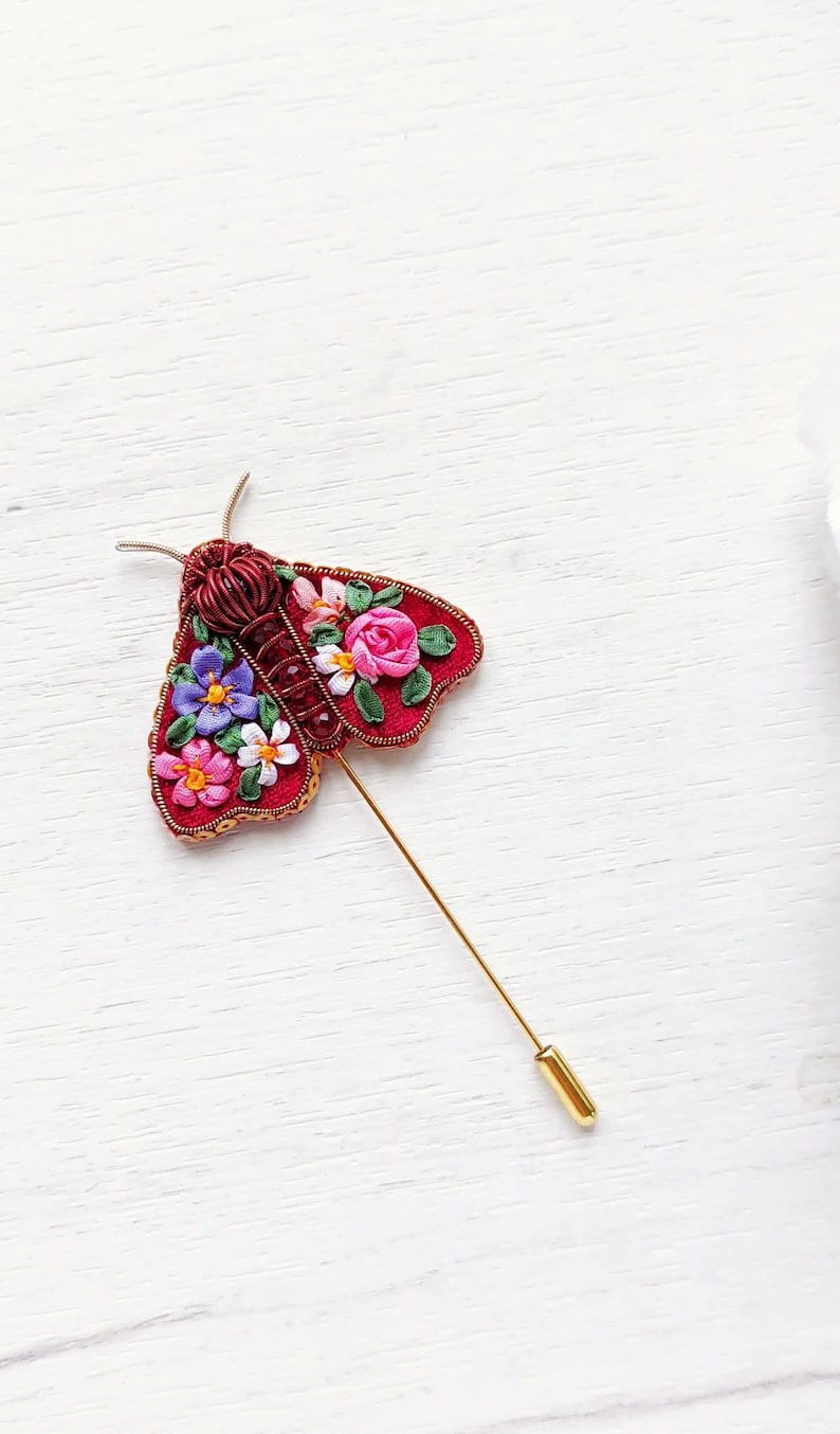 May include: A handcrafted red moth brooch with floral embroidery in pink, purple, and white. The brooch has a gold pin and is set against a white wooden background. The moth's body is a deep red, and the wings are adorned with colorful flowers.