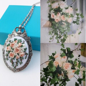 May include: A silver necklace with a white oval pendant featuring a delicate embroidered floral design of pink and white roses and green leaves.