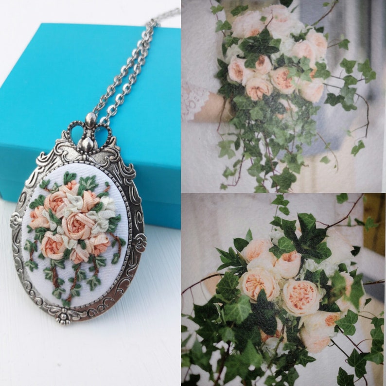 May include: A silver necklace with a white oval pendant featuring a delicate embroidered floral design of pink and white roses and green leaves.