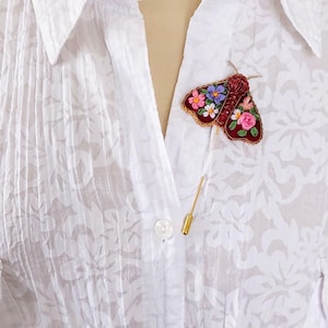 May include: A burgundy moth brooch with floral embroidery in pink, blue, and white is pinned to a white blouse. The brooch has a gold pin backing. The blouse has a floral pattern and is buttoned at the collar.
