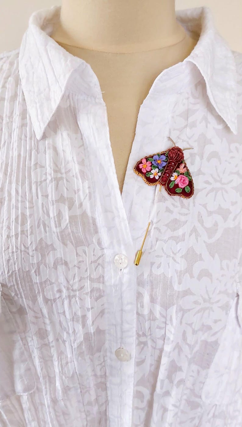 May include: A burgundy moth brooch with floral embroidery in pink, blue, and white is pinned to a white blouse. The brooch has a gold pin backing. The blouse has a floral pattern and is buttoned at the collar.