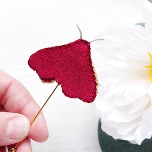 May include: A burgundy velvet butterfly pin with gold sequin trim and a gold pin backing. The butterfly is held in front of a white background with a white flower.