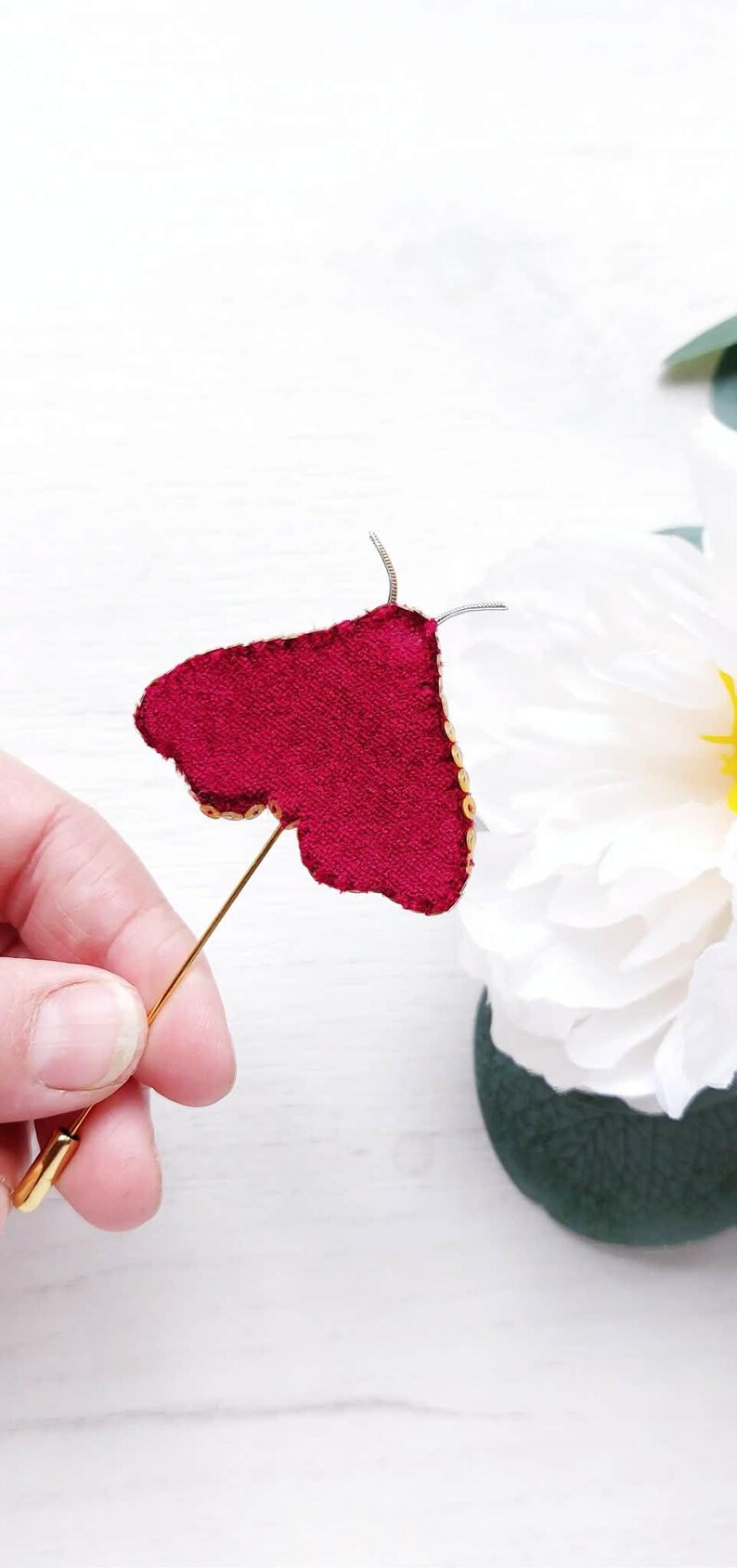May include: A burgundy velvet butterfly pin with gold sequin trim and a gold pin backing. The butterfly is held in front of a white background with a white flower.