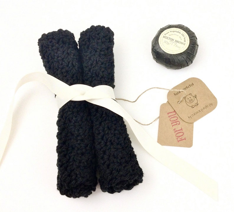 May include: Two black crocheted dishcloths tied together with a white ribbon and a brown gift tag that says "FOR YOU!" with a small pig illustration and the text "hand crocheted" in red. A round black soap bar with a white label that says "Pure Vegetable Oil Soap MOLTON BROWN 50g/1.75oz" is also included.