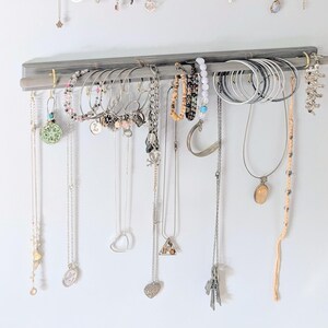 Puede incluir: Un organizador de joyas de madera gris que muestra varios collares, pulseras y pendientes. Las joyas incluyen piezas de plata, oro y cuentas. El organizador est&aacute; montado en una pared blanca, mostrando una colecci&oacute;n de accesorios.