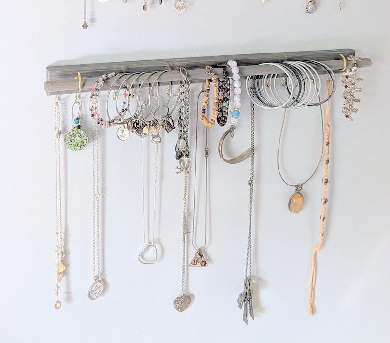 Puede incluir: Un organizador de joyas de madera gris que muestra varios collares, pulseras y pendientes. Las joyas incluyen piezas de plata, oro y cuentas. El organizador est&aacute; montado en una pared blanca, mostrando una colecci&oacute;n de accesorios.