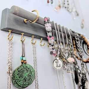 May include: A gray wooden jewelry organizer with gold-colored hooks and a rod. Various necklaces, bracelets, and earrings hang from the organizer. A beaded bracelet with pink and black beads is visible.