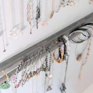 Puede incluir: Un organizador de joyas de madera gris montado en una pared, que muestra varios collares, pulseras y anillos. El organizador tiene ganchos para collares y una barra para pulseras y anillos. Las joyas son plateadas, doradas y con cuentas.