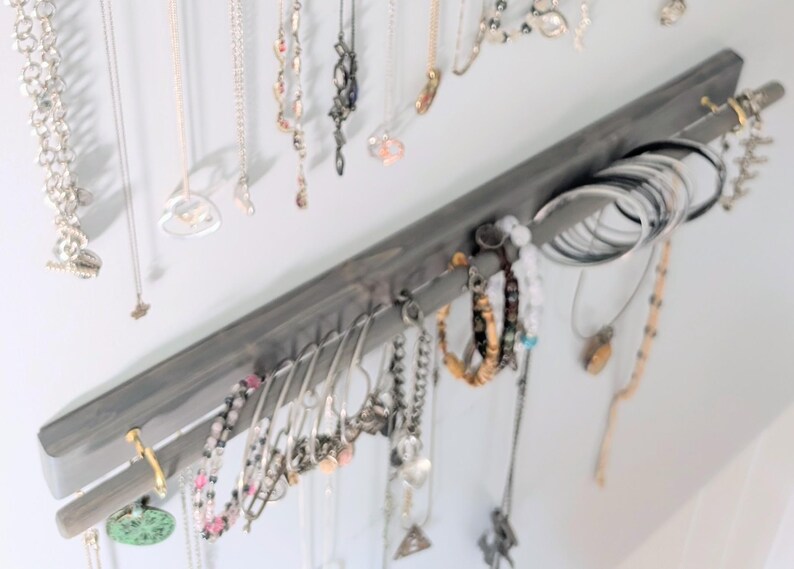 Puede incluir: Un organizador de joyas de madera gris montado en una pared, que muestra varios collares, pulseras y anillos. El organizador tiene ganchos para collares y una barra para pulseras y anillos. Las joyas son plateadas, doradas y con cuentas.