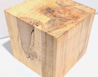 Puede incluir: Un cubo de madera maciza con un patrón de veta natural en tonos beige y marrón claro. La superficie muestra intrincados patrones de remolinos, marcas orgánicas de color marrón oscuro y una textura de madera visible en sus caras.