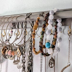 Puede incluir: Una exhibici&oacute;n de varios collares y pulseras. La joyer&iacute;a incluye cadenas de plata, pulseras de cuentas con cuentas turquesas y marmoladas, y collares con colgantes florales y geom&eacute;tricos. La joyer&iacute;a cuelga de un estante de madera.