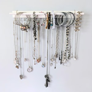 May include: A collection of necklaces, bracelets, and earrings hang from a white wooden jewelry organizer. The jewelry includes silver and gold chains, beaded necklaces, and various charms. Bangles and hoop earrings are also visible.