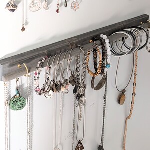 Puede incluir: Un organizador de joyas de madera gris con m&uacute;ltiples ganchos y una barra, que muestra varios collares, pulseras y aretes. Las joyas incluyen piezas de plata, oro y cuentas, creando una soluci&oacute;n de almacenamiento elegante.
