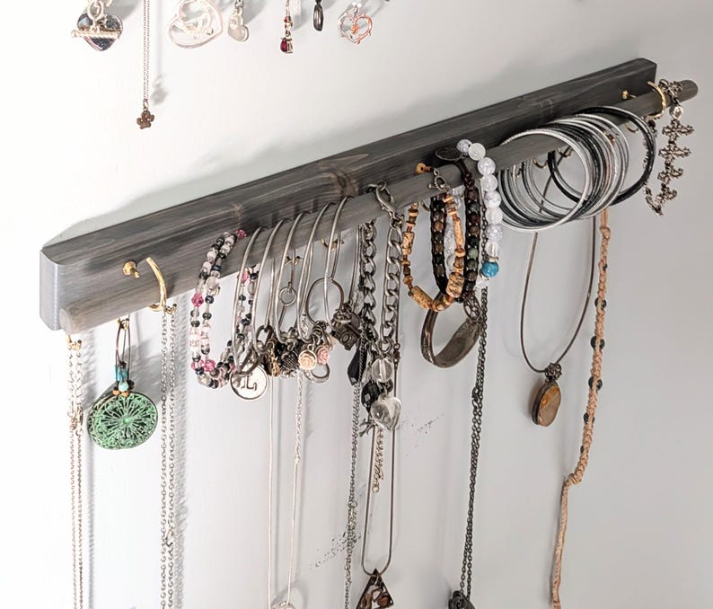 Puede incluir: Un organizador de joyas de madera gris con m&uacute;ltiples ganchos y una barra, que muestra varios collares, pulseras y aretes. Las joyas incluyen piezas de plata, oro y cuentas, creando una soluci&oacute;n de almacenamiento elegante.