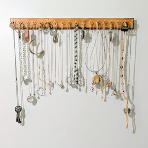 May include: A rectangular wooden wall-mounted jewelry organizer featuring a horizontal row of metal hooks. Numerous necklaces, chains, and pendants of varying lengths, colors, and styles hang from the hooks, creating a display of accessories.