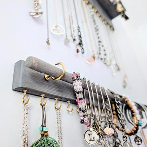 Puede incluir: Un organizador de joyas de madera gris con m&uacute;ltiples collares, pulseras y pendientes colgando. El organizador est&aacute; montado en una pared blanca y presenta ganchos dorados y una barra curva para colgar joyas.